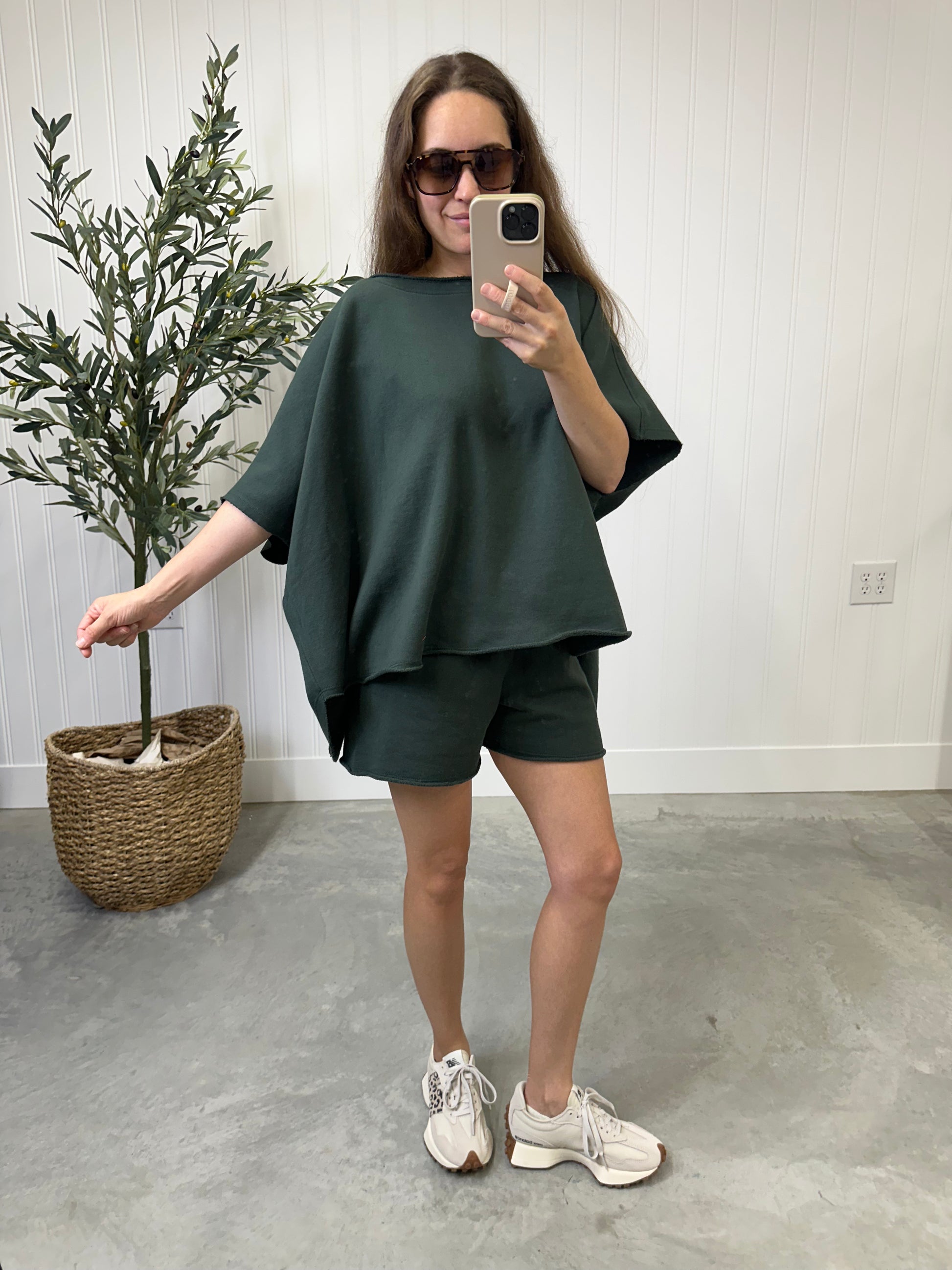 Woman taking a mirror selfie wearing a dark green outfit with shorts and white sneakers in a room with a plant.