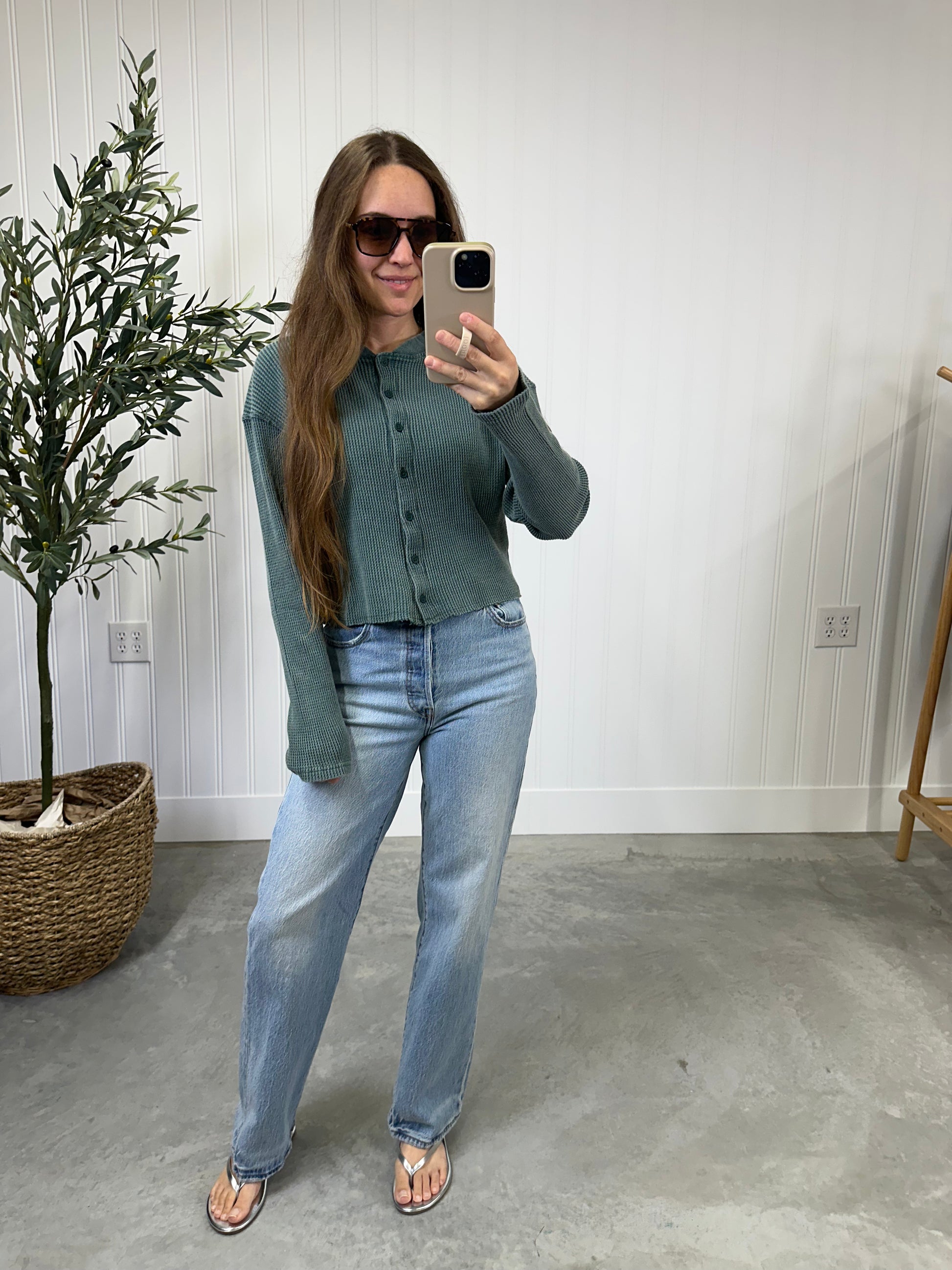 Woman taking a mirror selfie wearing a green shirt and blue jeans in a room with a plant and light-colored walls. Evergreen Waffle Top