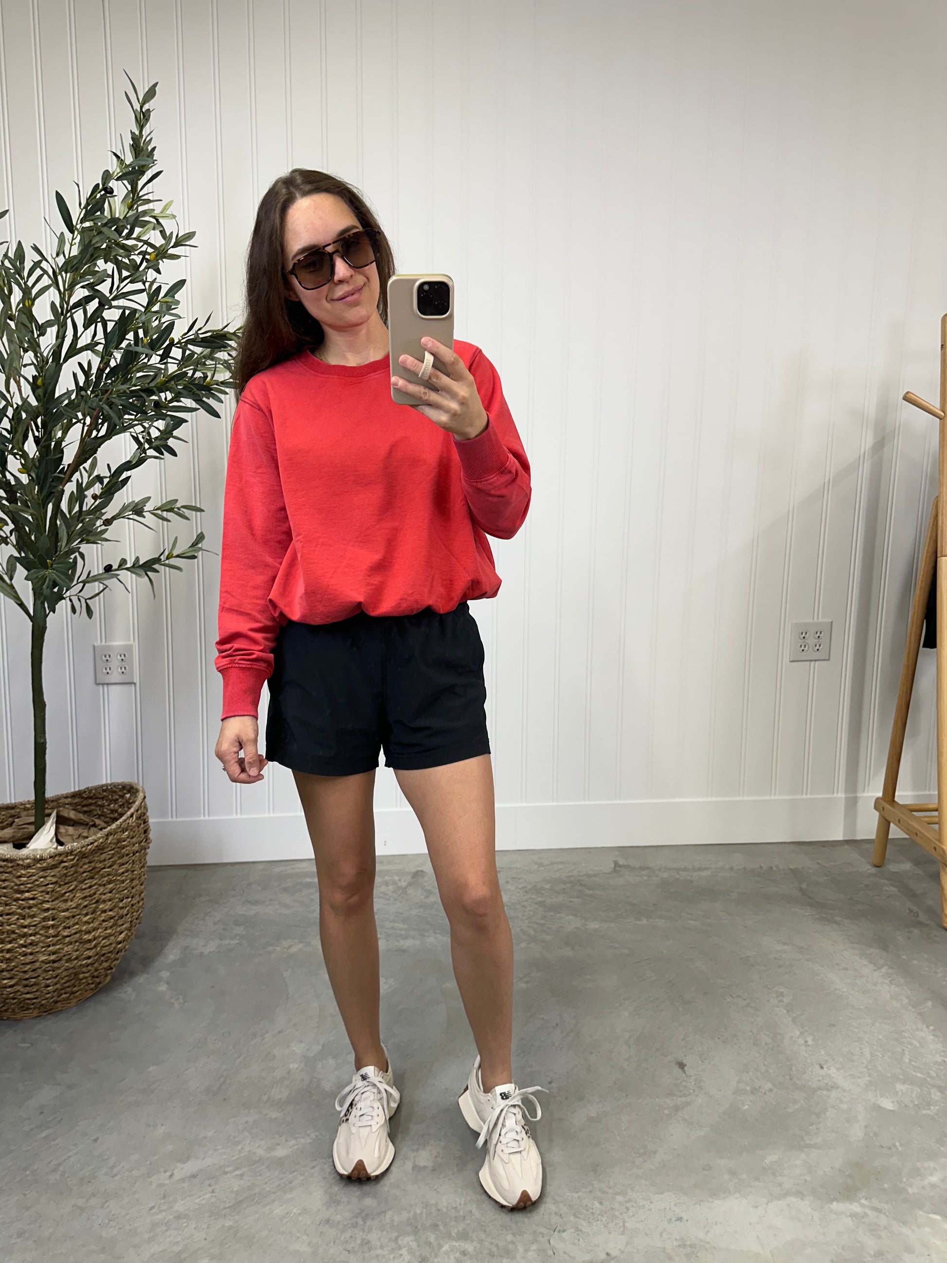 Woman taking a mirror selfie wearing a red sweatshirt, black shorts, and white sneakers in a room with a plant and wooden stool. Glazed Cherry Bubble Pullover