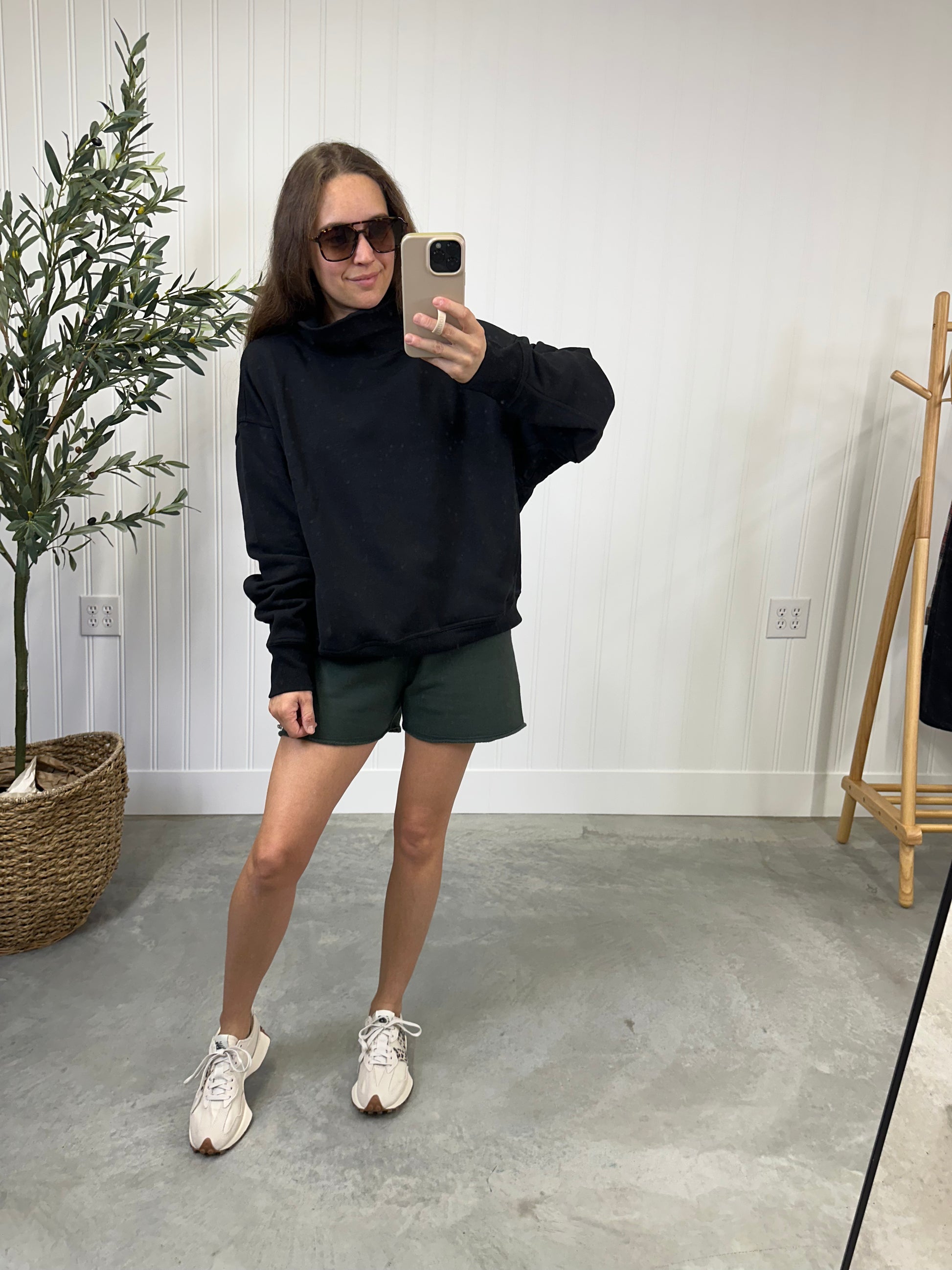 Person taking a mirror selfie wearing a black hoodie, green shorts, and white sneakers in a room with a plant and coat rack.