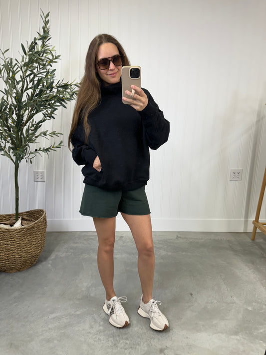 Person taking a mirror selfie wearing a black sweatshirt, green shorts, and white sneakers in a room with a plant and light-colored wall.