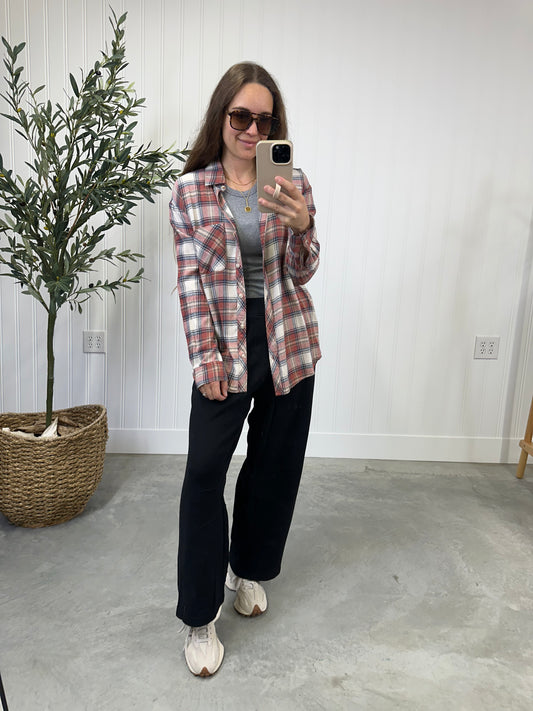 Woman taking a mirror selfie wearing a plaid shirt, gray top, and black pants in a room with a plant and light-colored walls.