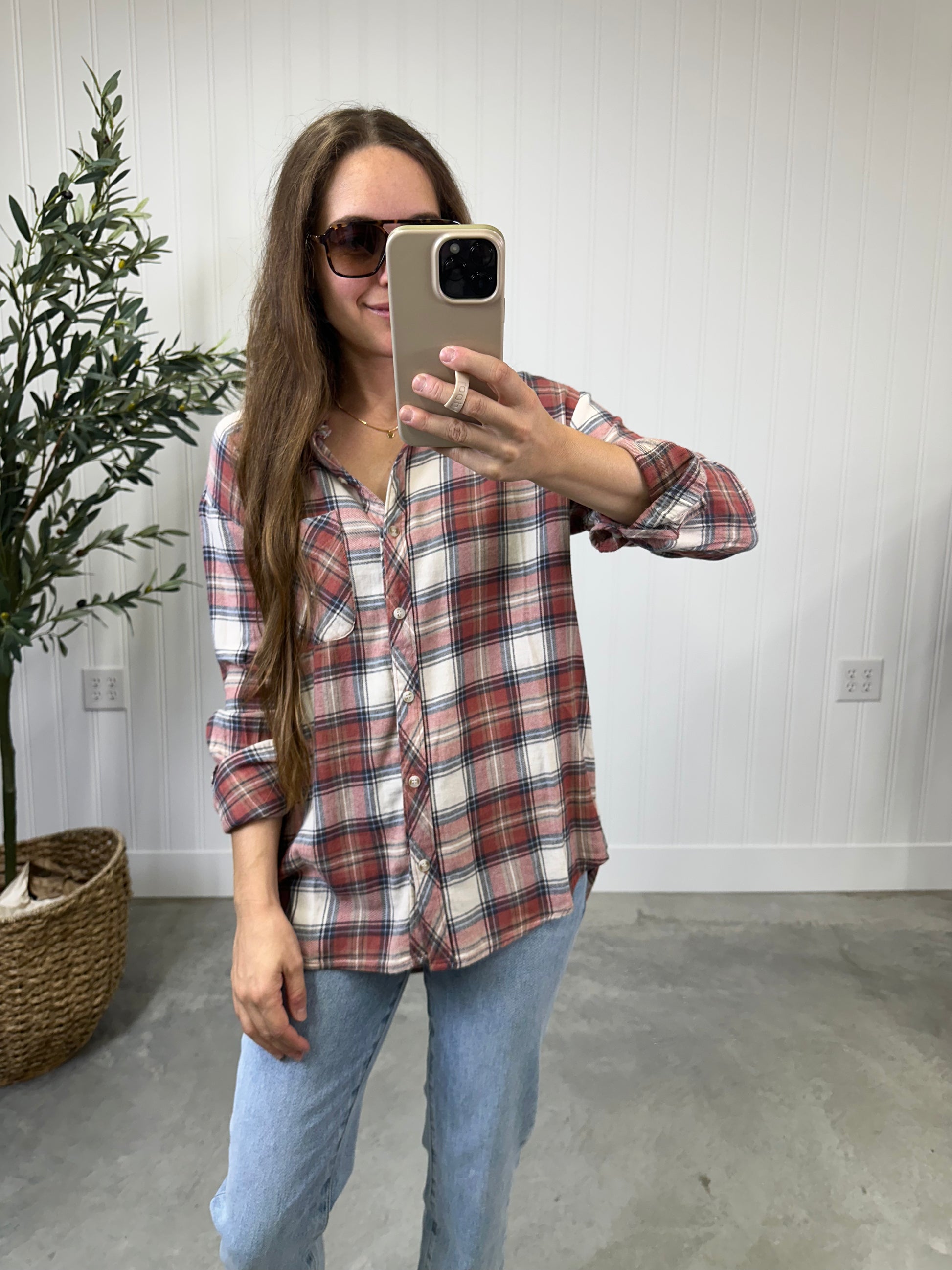 Woman taking a mirror selfie wearing a plaid shirt and jeans in a room with a plant and light-colored wall.