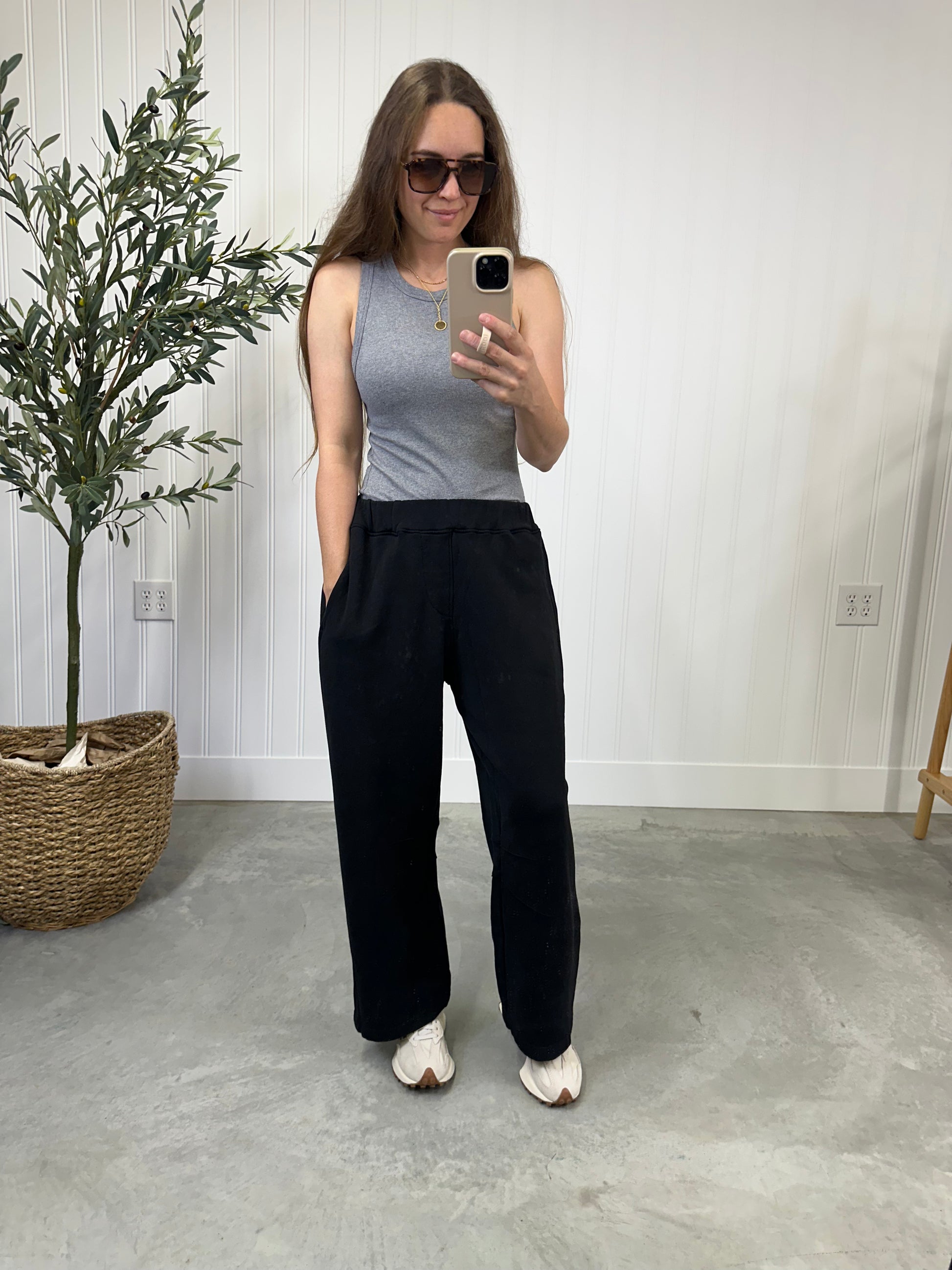Woman taking a mirror selfie wearing a gray tank top and black pants in a room with a plant and light-colored floor. The Onyx Barrel Lounge Pants